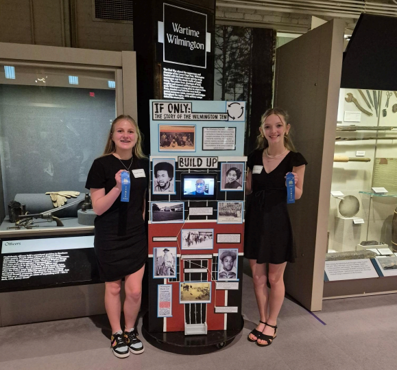 two students stand in front of an exhibit that they created while holding blue ribbons