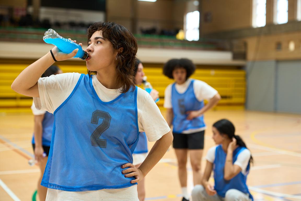 A young female basketball player drinks a blue sports drink from a bottle during practice.