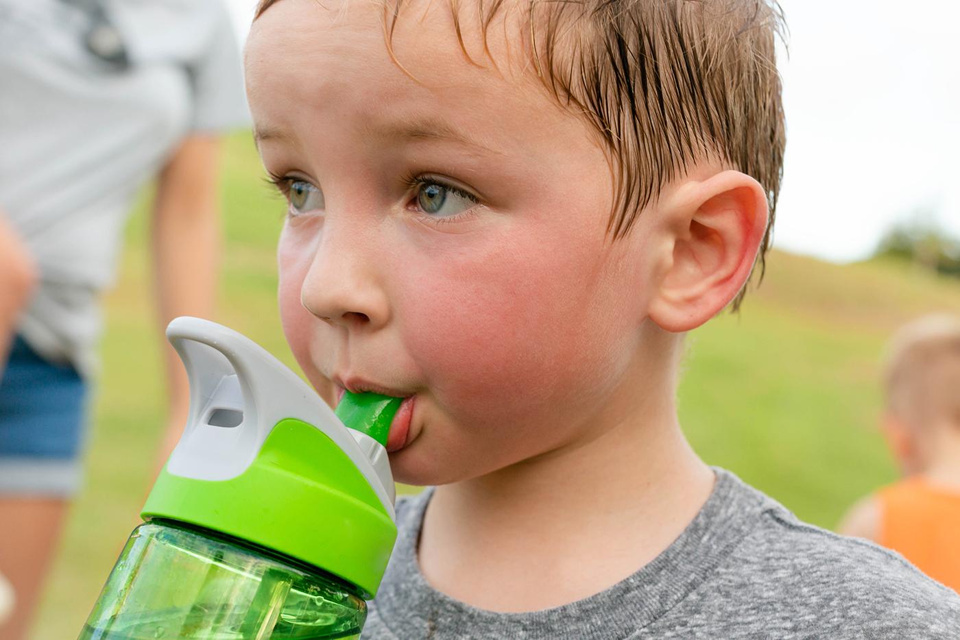 A young child with a flushed face due to the heat drinks from a water bottle.