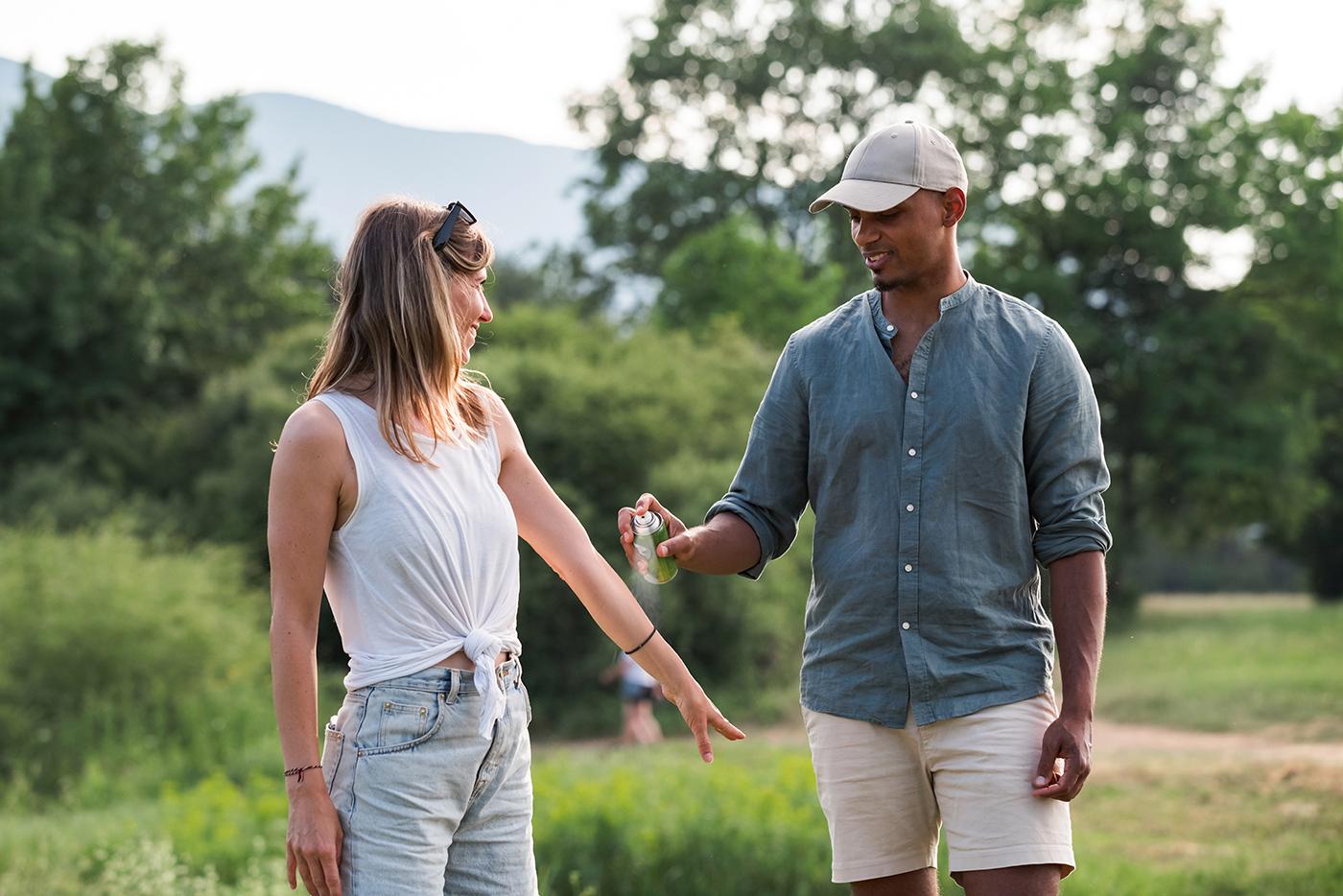 A young couple sprays one another with insect repellent before an outdoor hike.