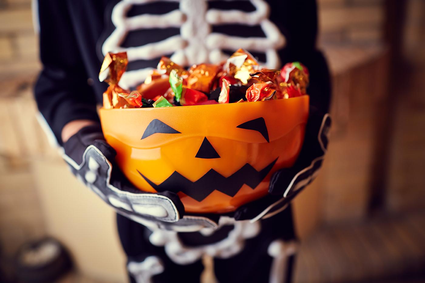 A child in a skeleton costume holds a Halloween-themed container full of candy.