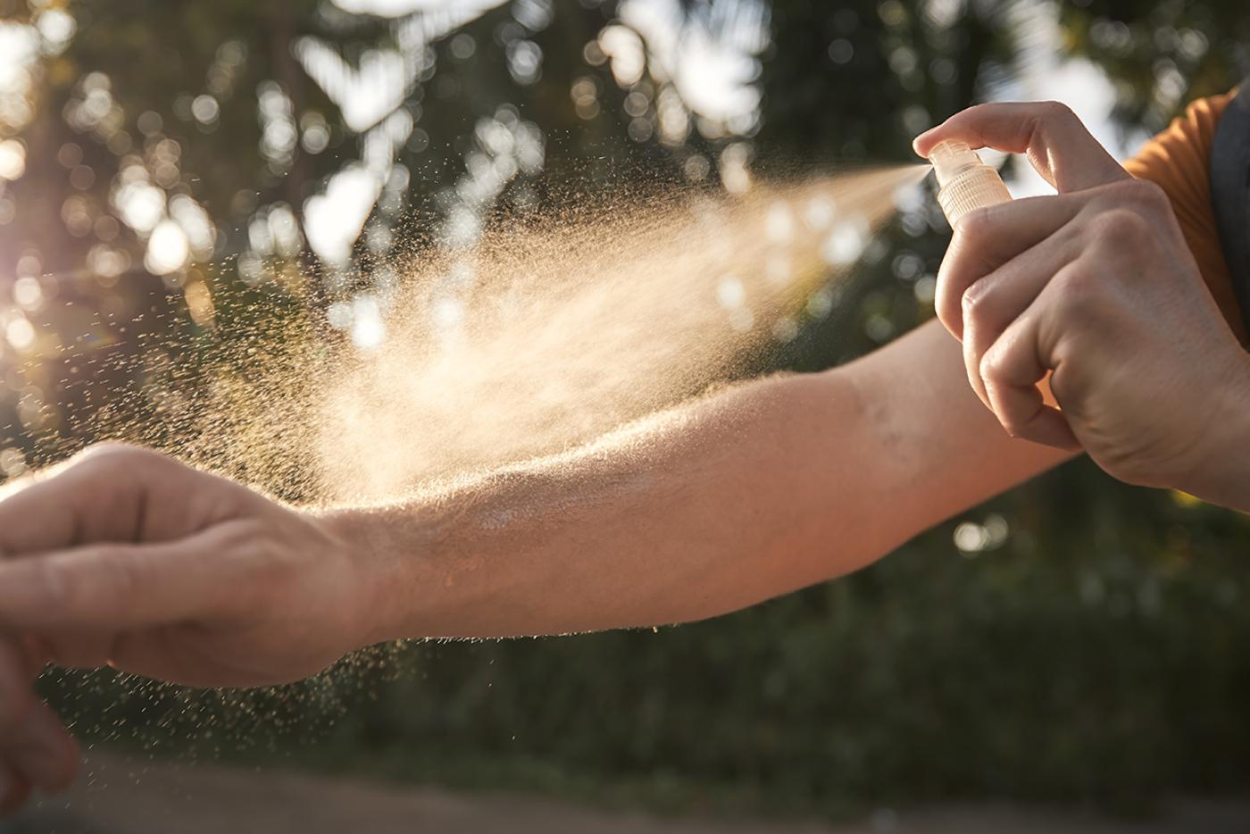 A person holds mosquito repellent in one hand while spraying the mist on their other arm.