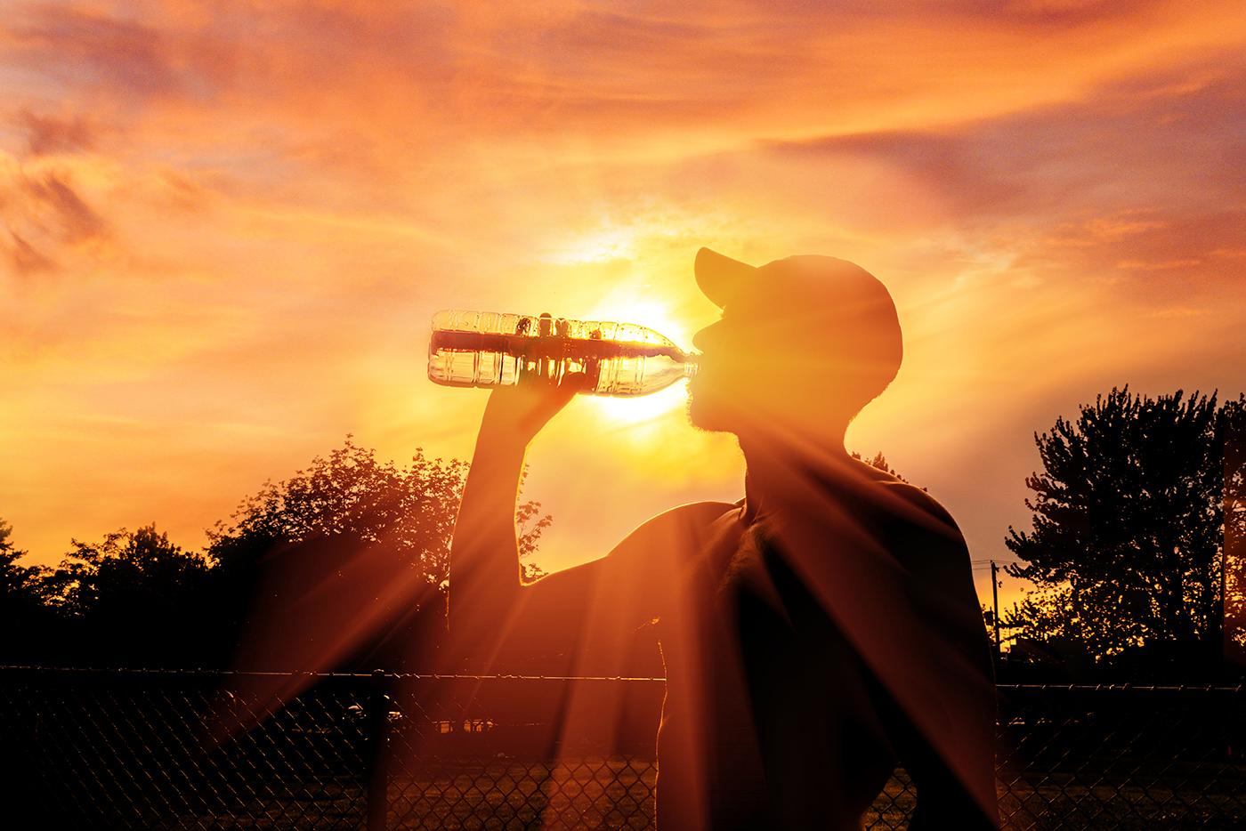 A man drinks from a bottle of water while standing outside during a heat wave. The sun shines behind him.