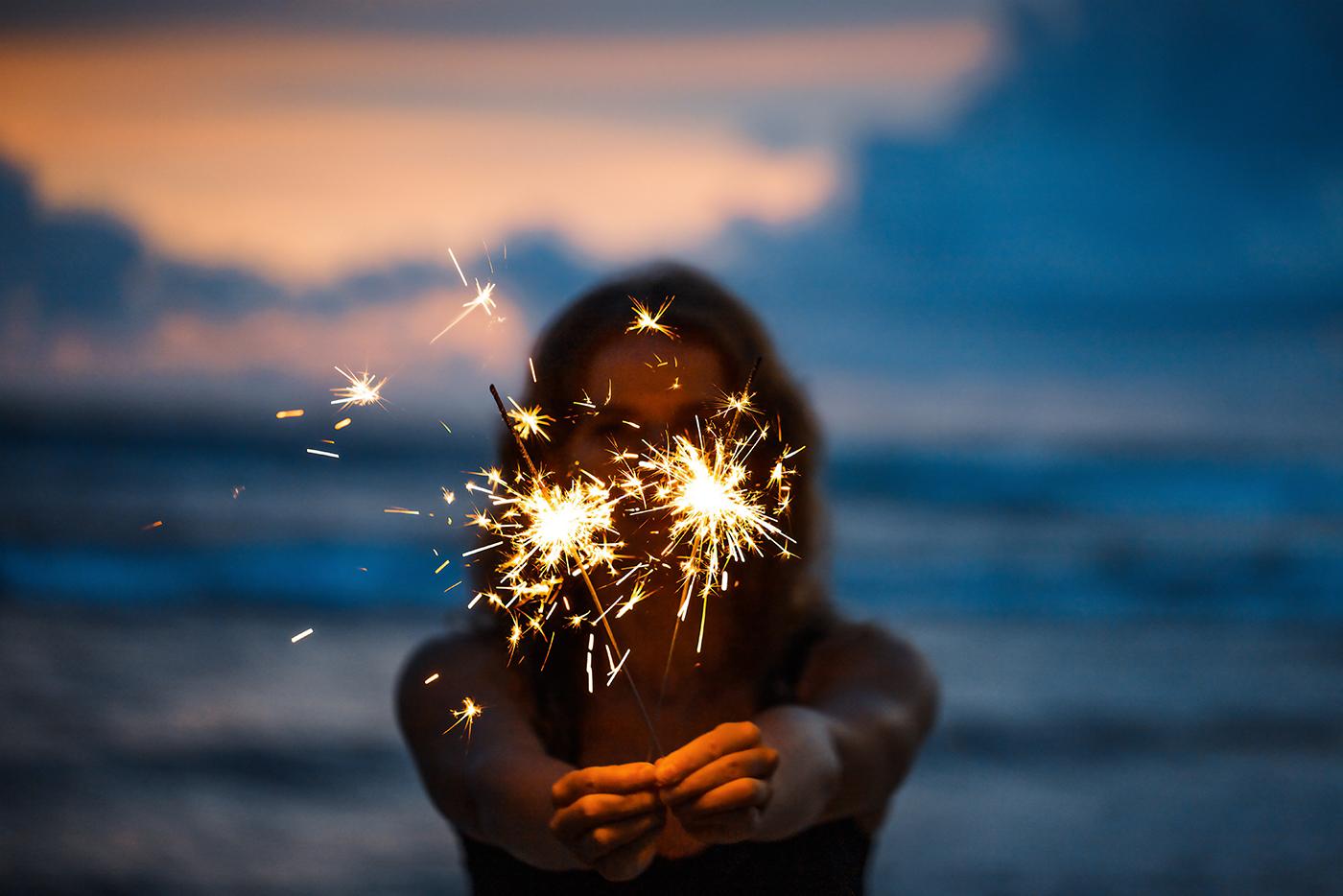 A woman holds two lit sparklers in her hands while standing on the beach.