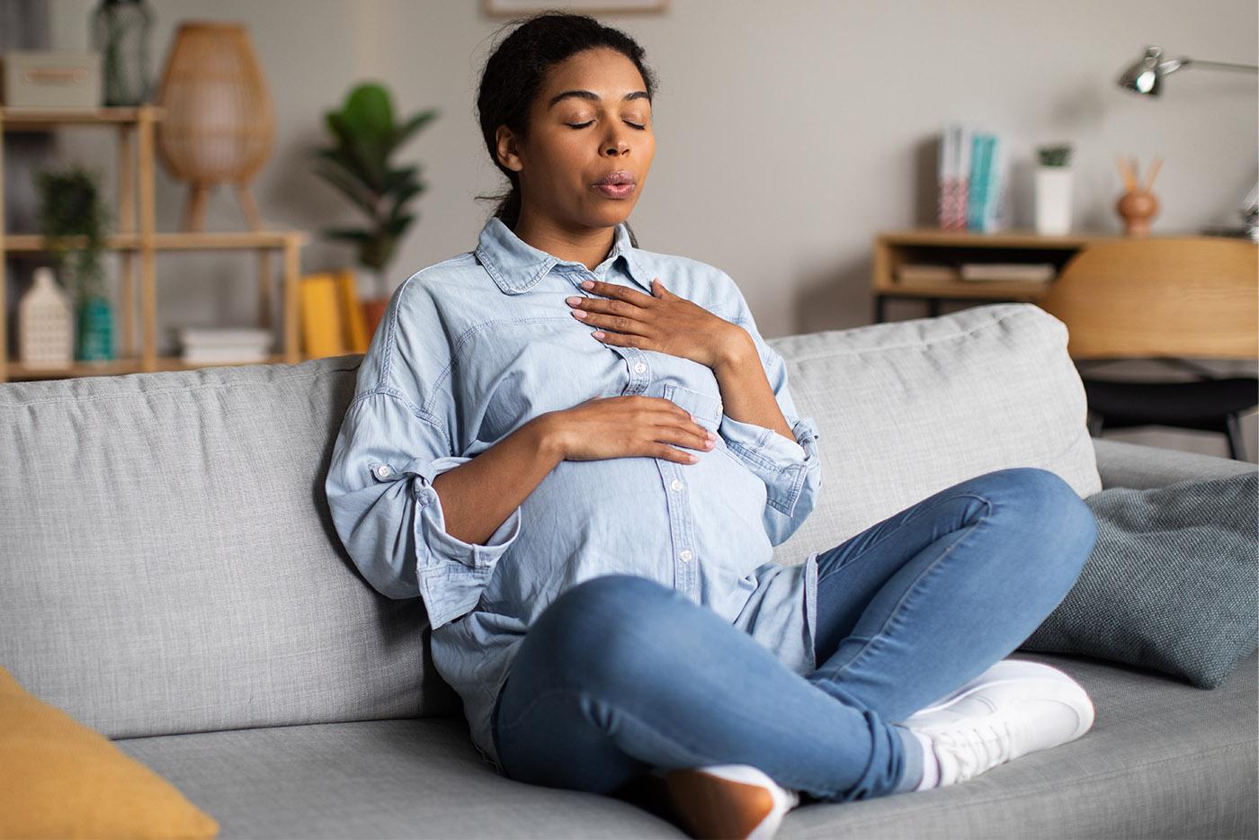 A pregnant woman sitting on a couch rests her hands on her heart and stomach.