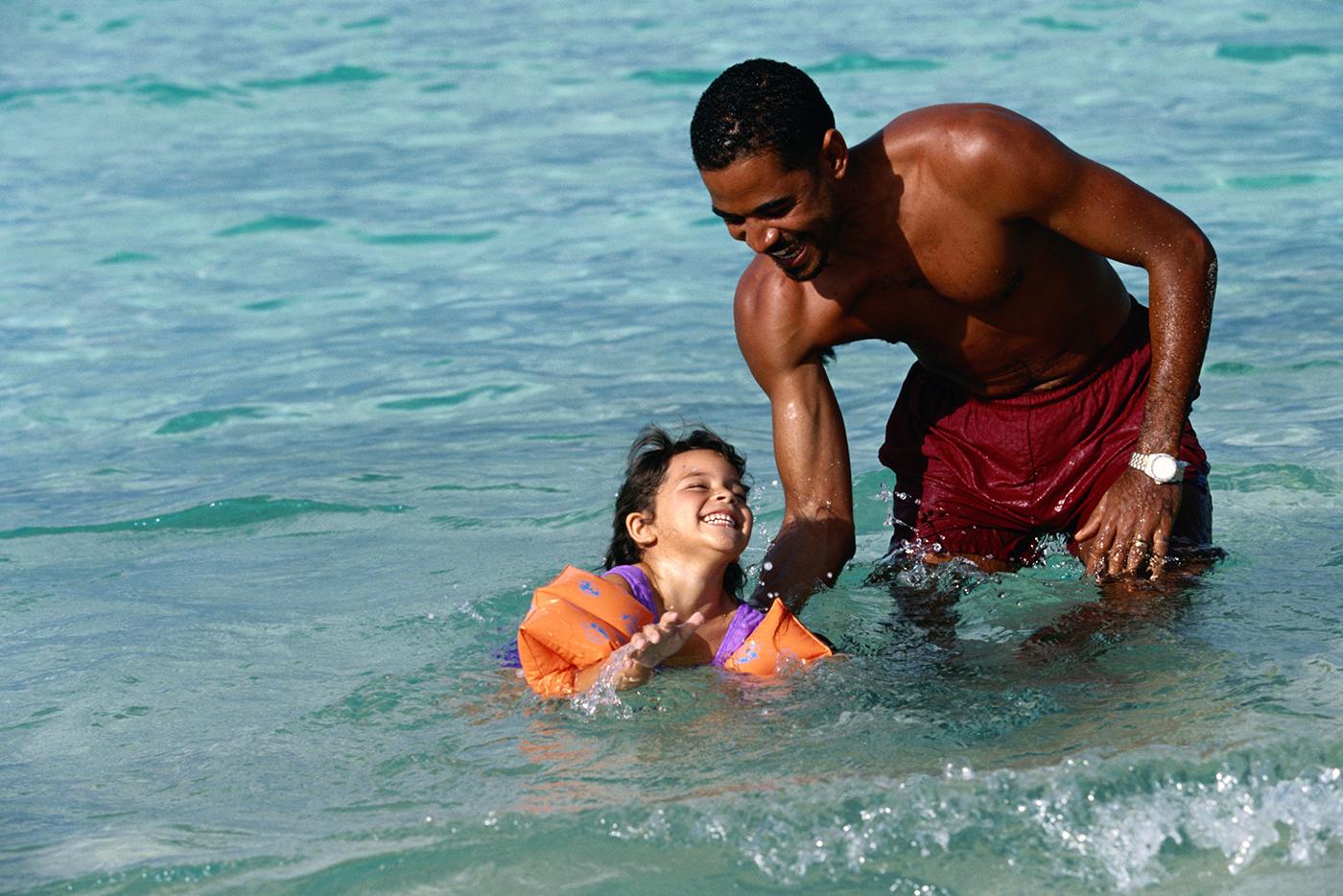 A father helps his daughter as she swims in the ocean with arm floaties.