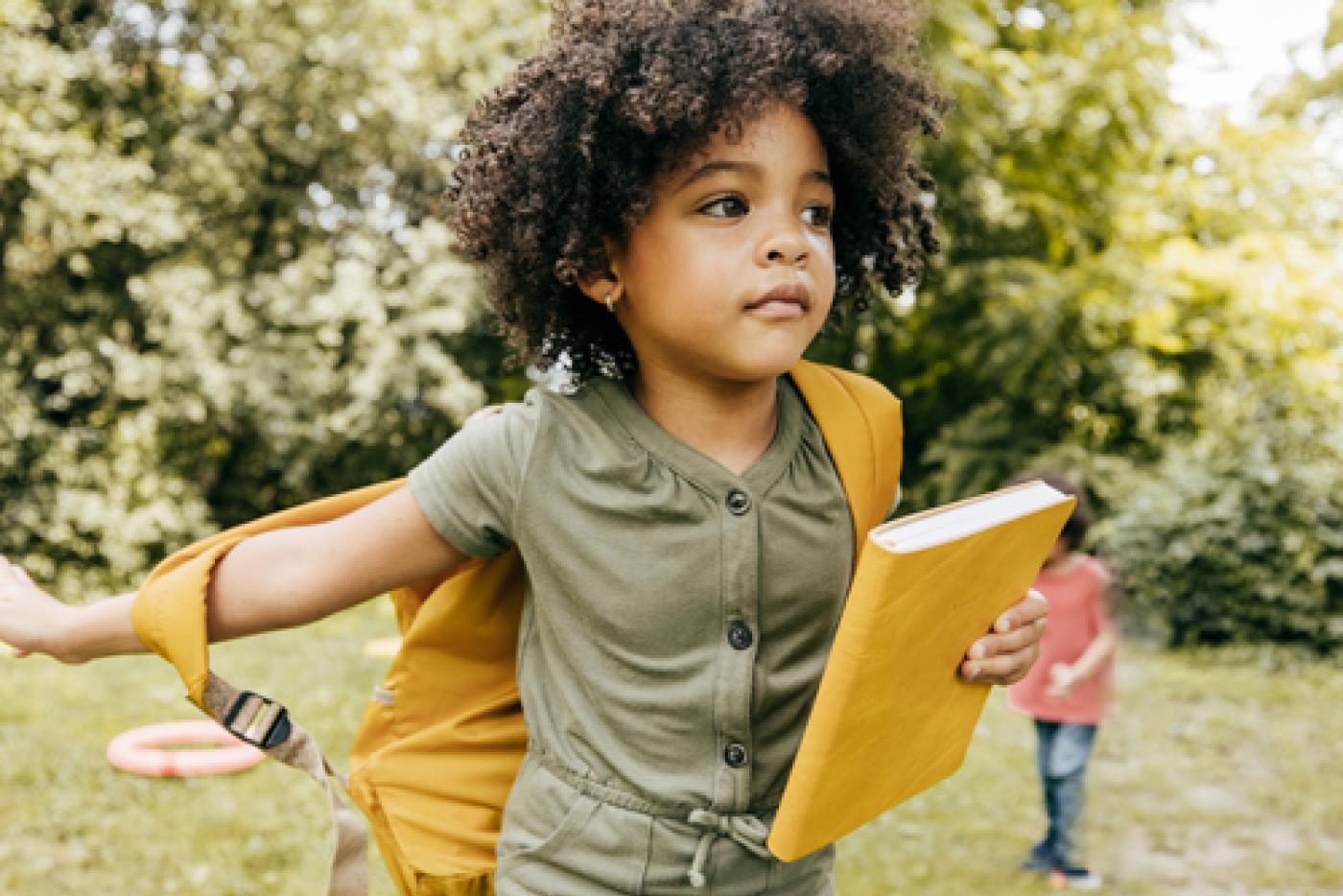 A young child places their arm through their backpack and holds a notebook.