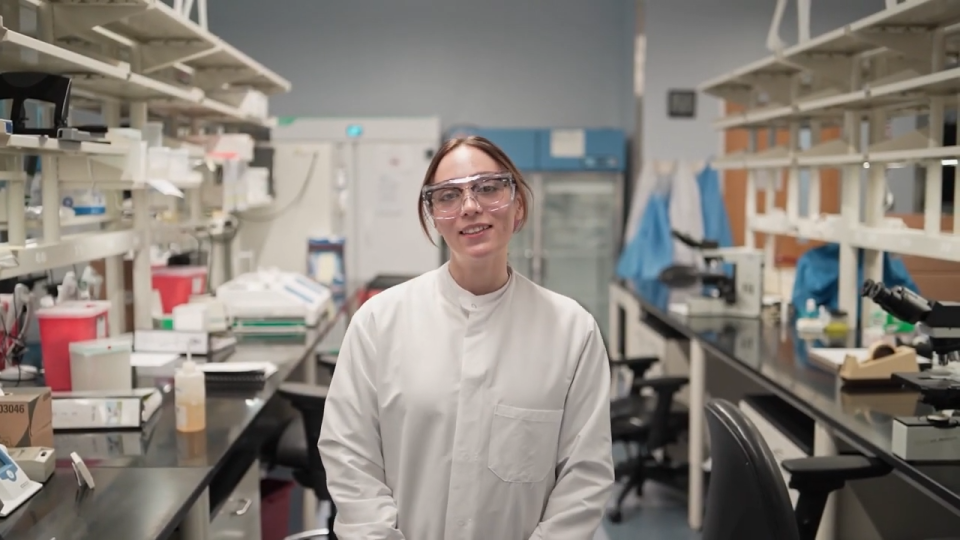 Kaila Kurtz stands in the Environmental Microbiology Lab, with equipment behind her. She's wearing protective eye gear and a lab coat.