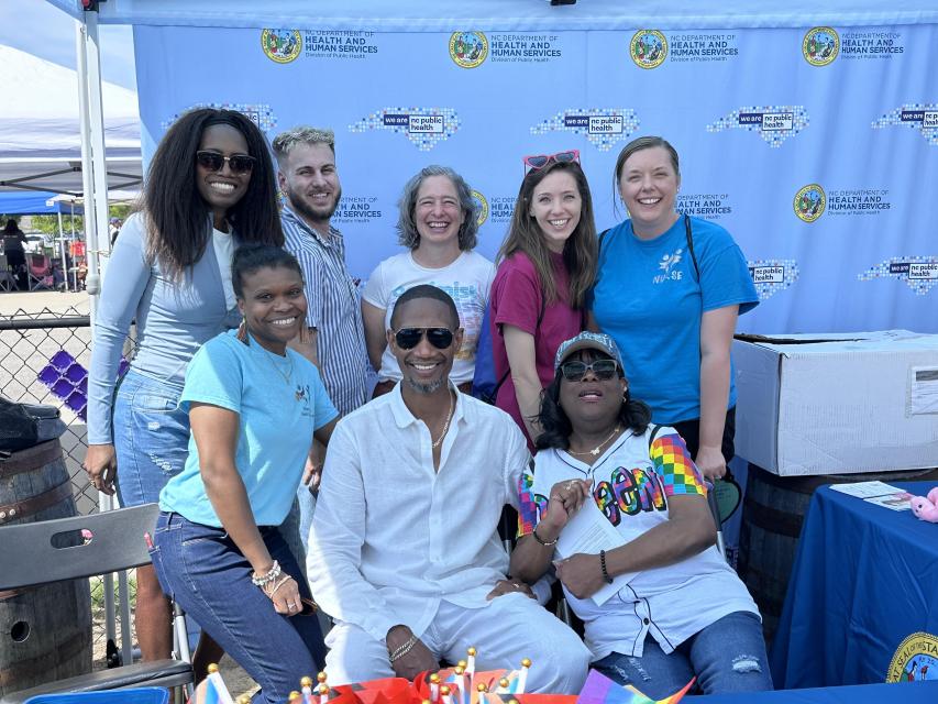 NCDHHS and local health department staff at an outdoor community event. Three are seated and five are standing against a blue backdrop with logos.