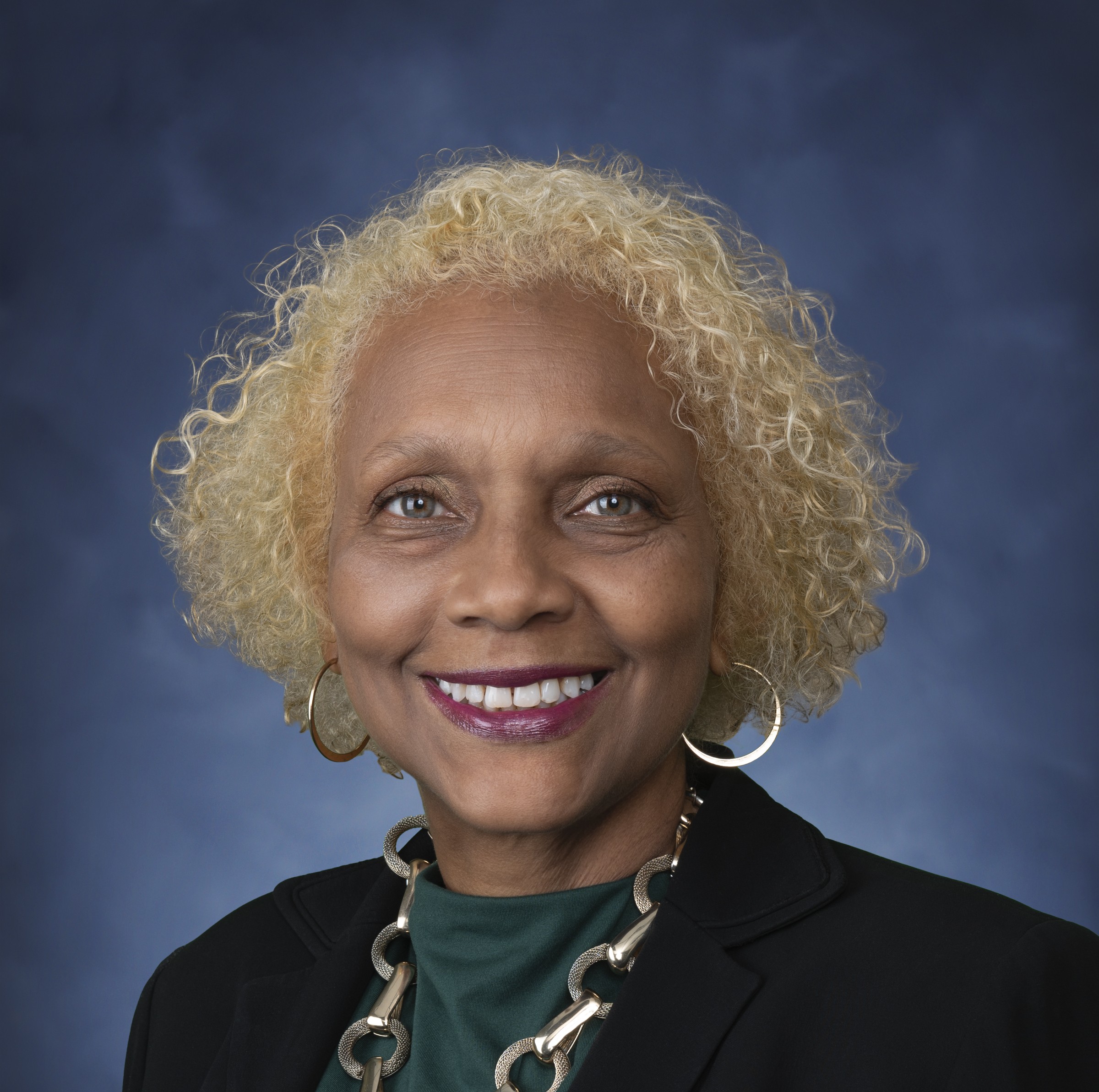 Portrait of Director of the Office of Victim Compensation Services Diane Barber in a black suit and green blouse standing in front of a blue backdrop