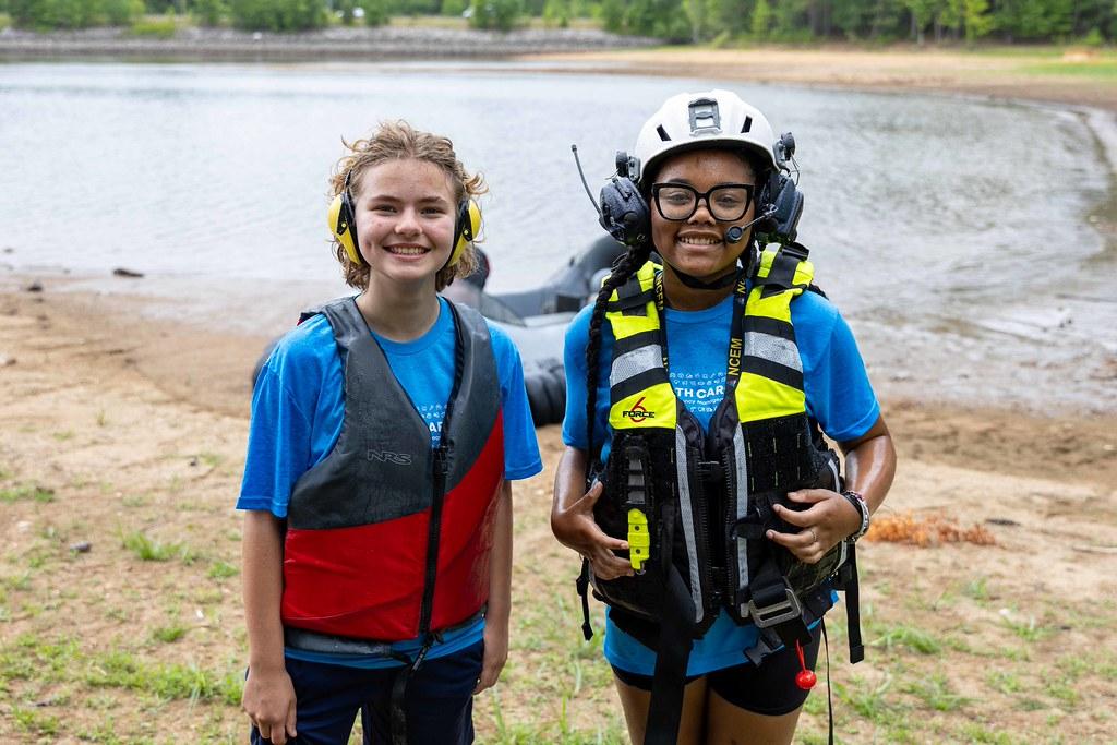Two young women stand by a lake in water rescue gear.