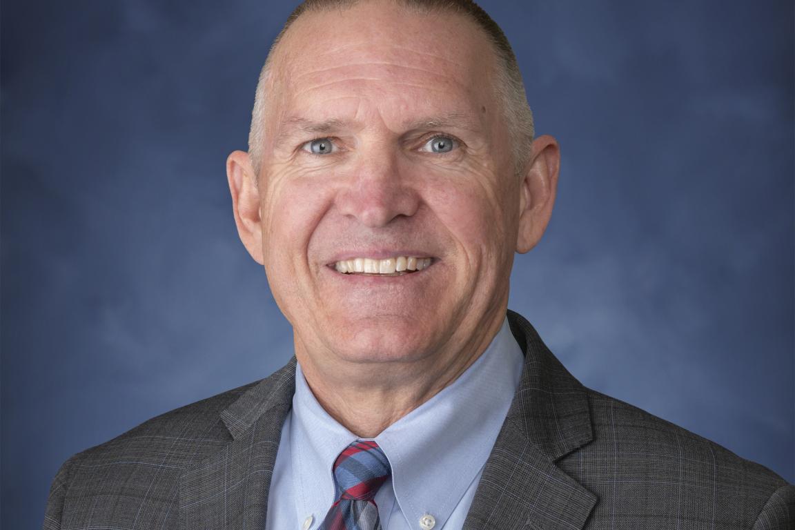 Portrait of Jeffrey Smythe. White male in a grey suit in front of blue background.