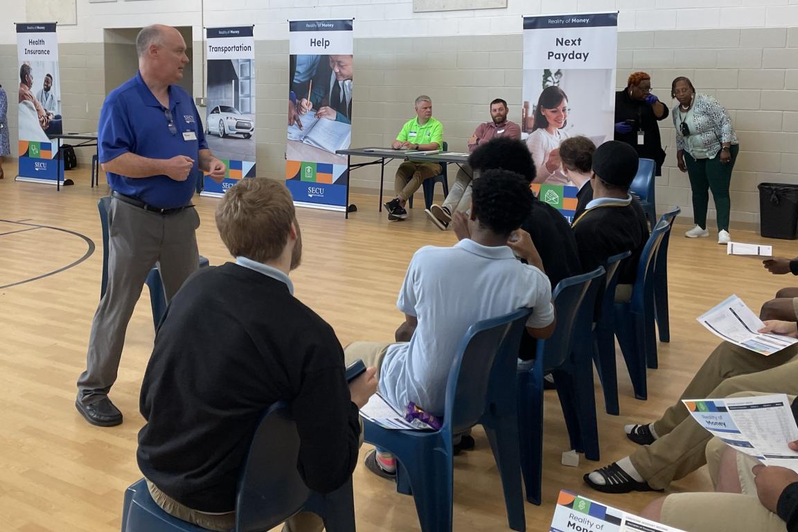 A person stands addressing a seated group youth in a gymnasium.