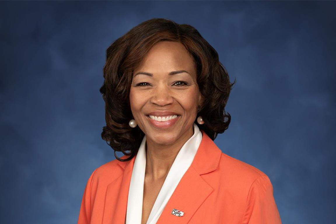 Black woman with orange suite jacket and white blouse.