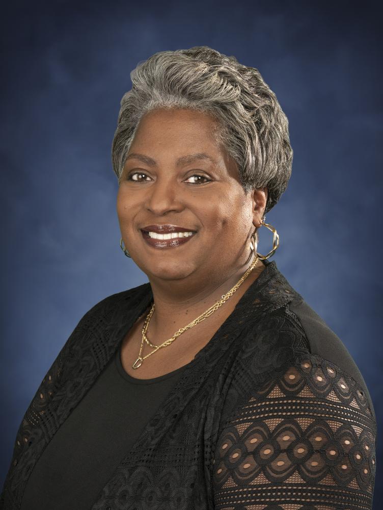 Sidra Owens (Black female with short grey hair) in black top in front of blue background.