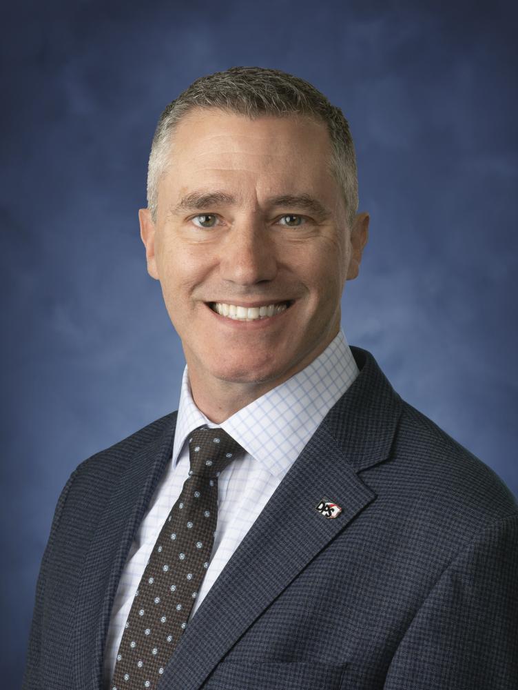 White male wearing grey suit, with white button-up shirt and spotted grey tie.
