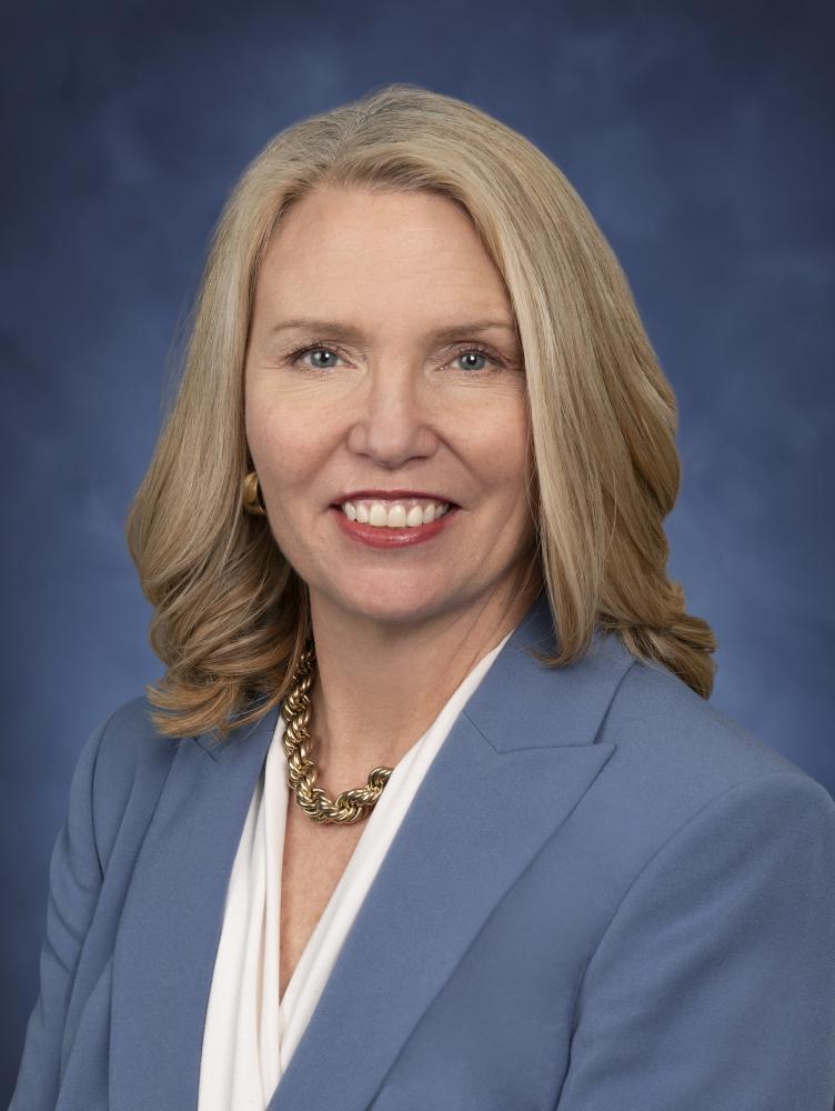 Profile photo of Caroline Valand,  (white female with medium length blonde hair) in a white blouse with a blue blazer in front of blue background.