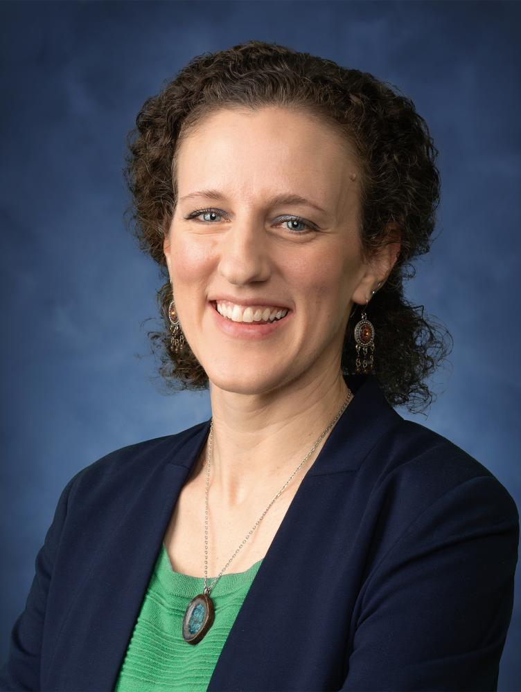 White woman the short brown hair tied back. She is wearing a green shirt and navy blazer.