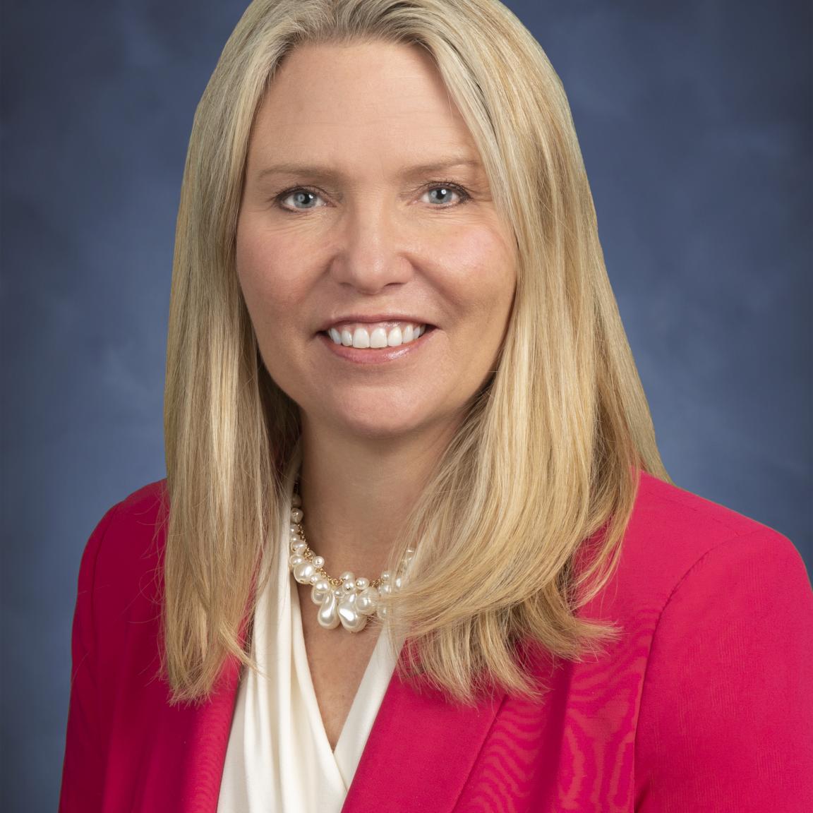 Caroline Valand (white female with medium length blonde hair) in pink blazer in front of blue background.