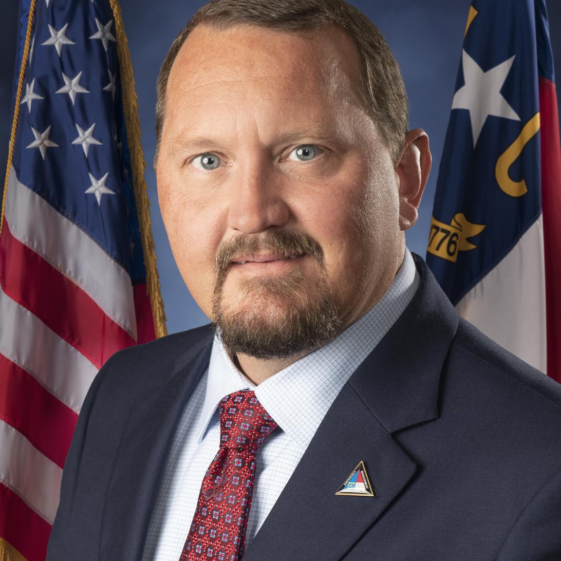 White male wearing a navy suit, white button-up shirt and red tie posed in front of US and NC flags.