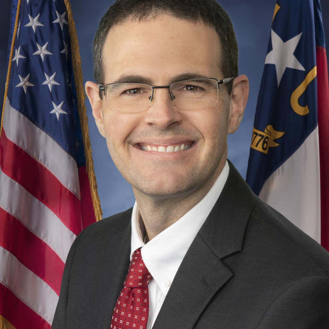 White male with glasses wearing a black suit, white button-up shirt and red tie posed in front of US and NC flags.