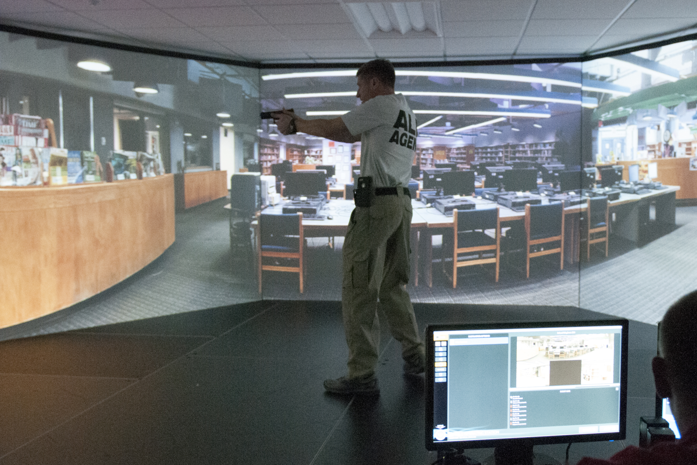 Male standing in middle of a room in front of a large screen displaying what appears to be a library. Man has a gun drawn.