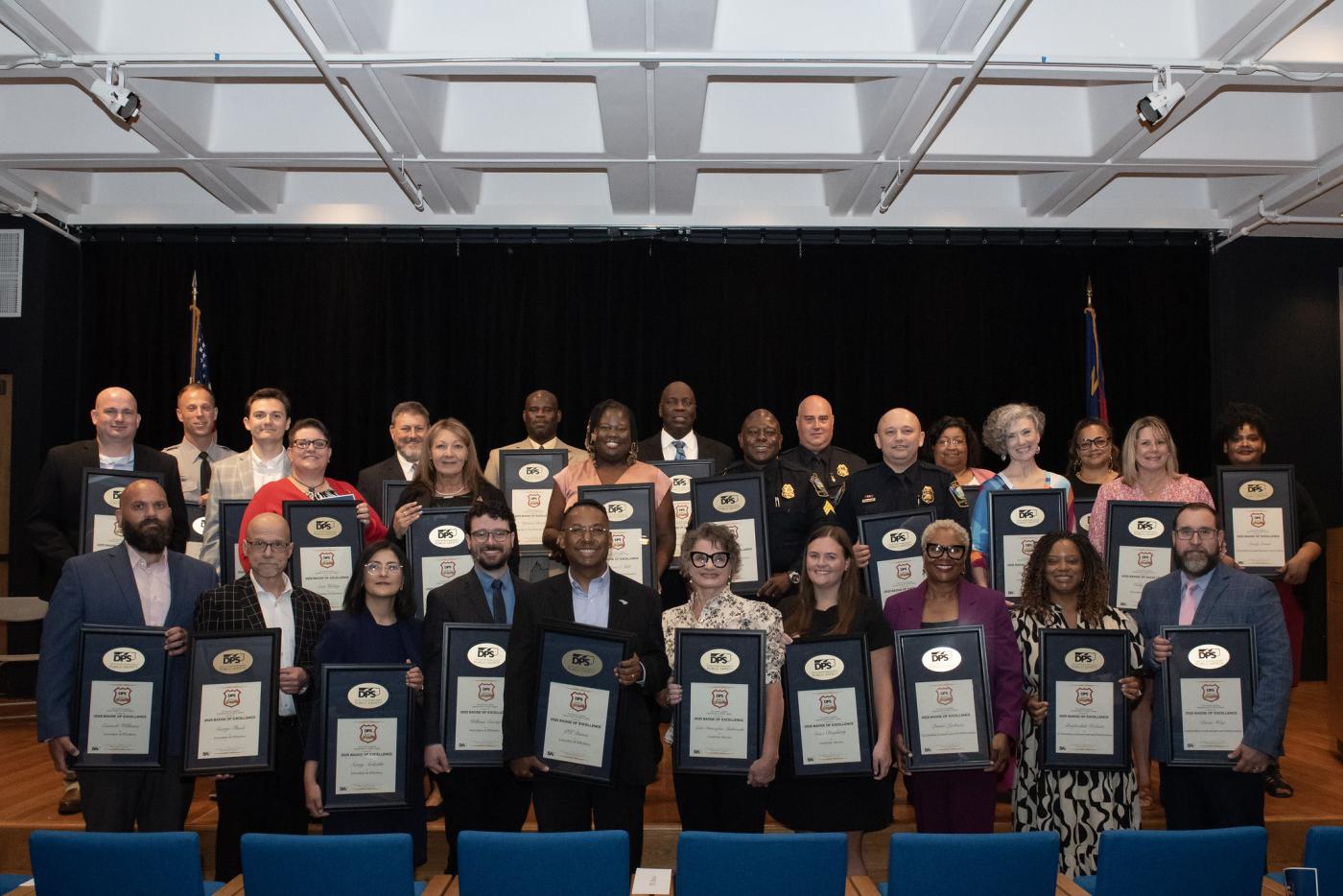 Multiple people pose together in an auditorium. Everyone holds a plaque indicating the award they are receiving.