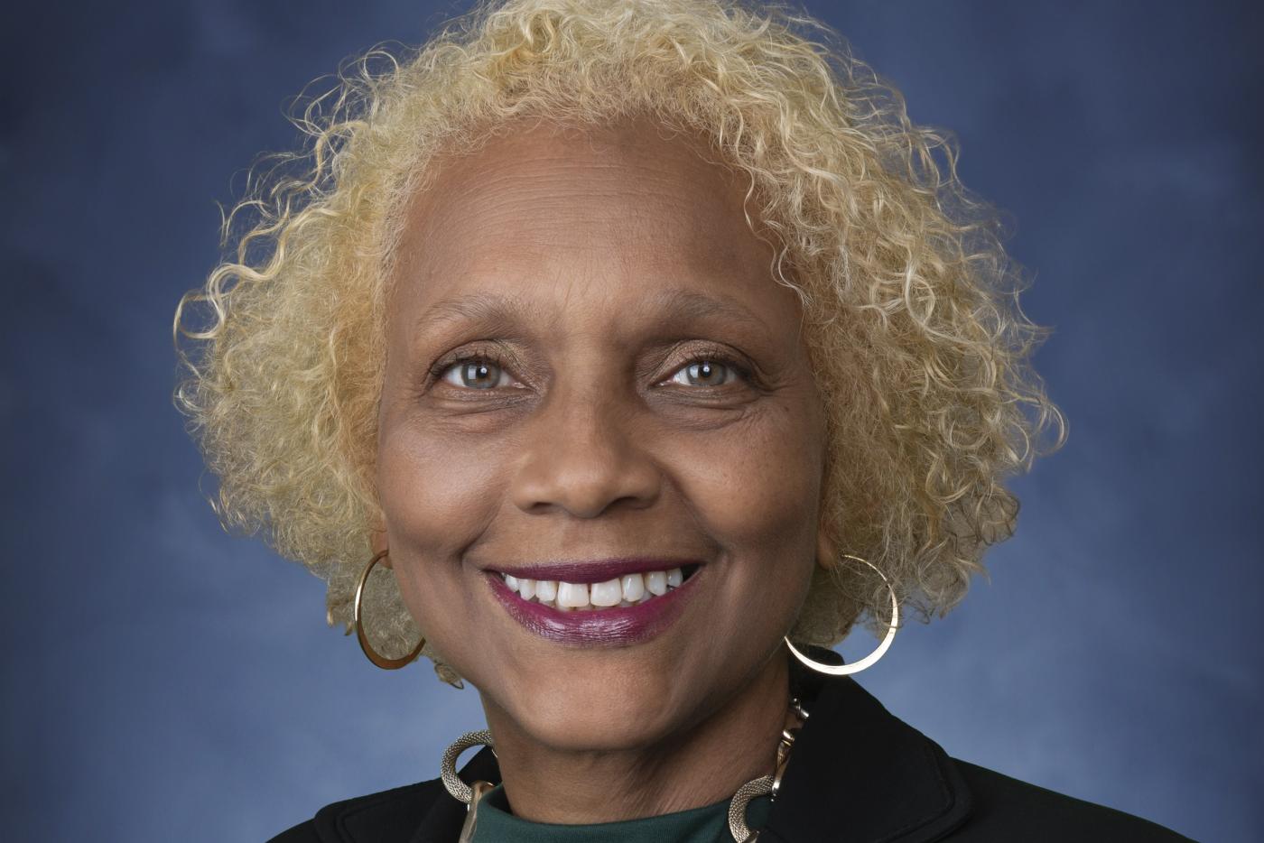 Portrait of Director of the Office of Victim Compensation Services Diane Barber in a black suit and green blouse standing in front of a blue backdrop