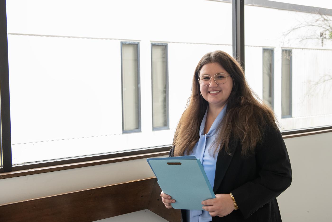 Female professionally dressed holding a folder and standing in front of a window