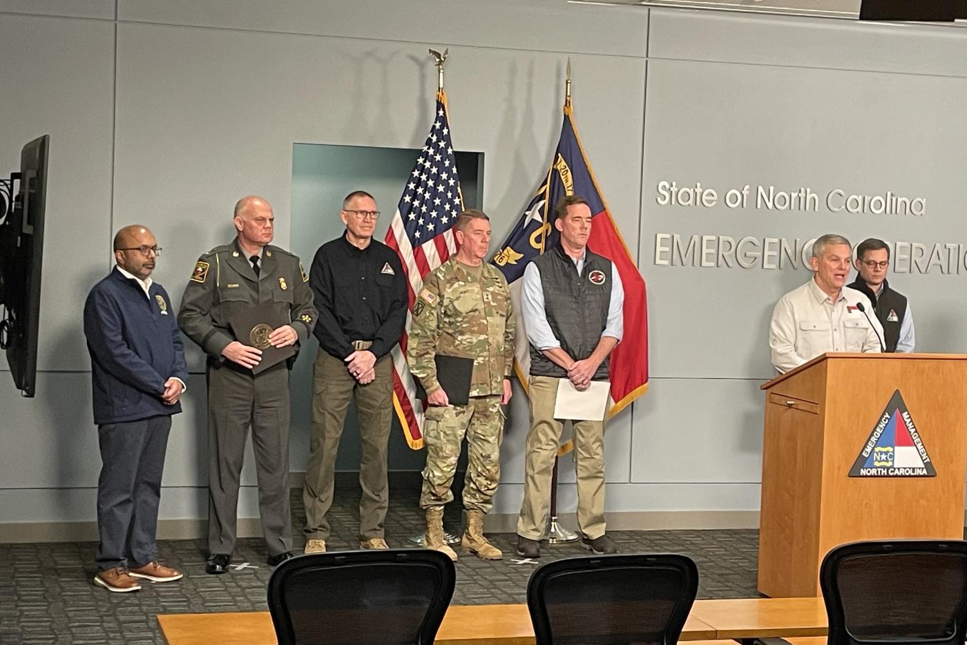 Five men stand on stage. One man, Gov. Stein, stands at podium to the right. Behind him stands another man.