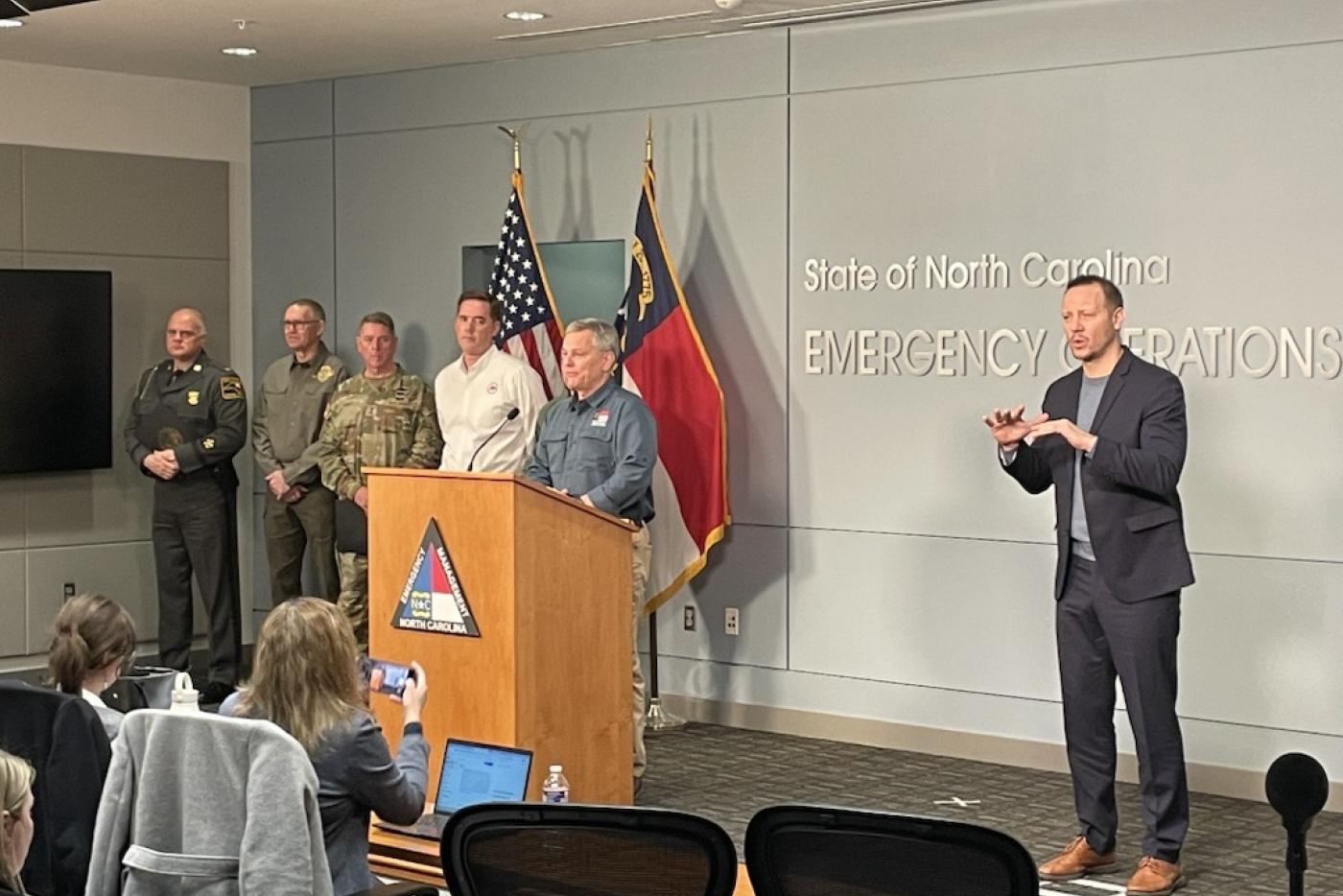 Six men stand on stage. One man, Gov. Stein, stands at podium at the center.