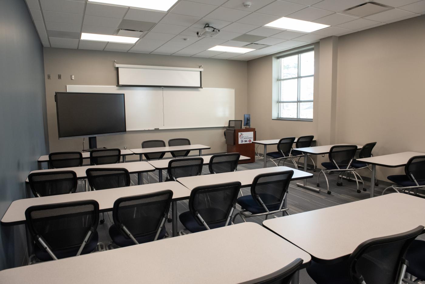 Room with whiteboard and TV at front. Four rows of tables with chairs at each.