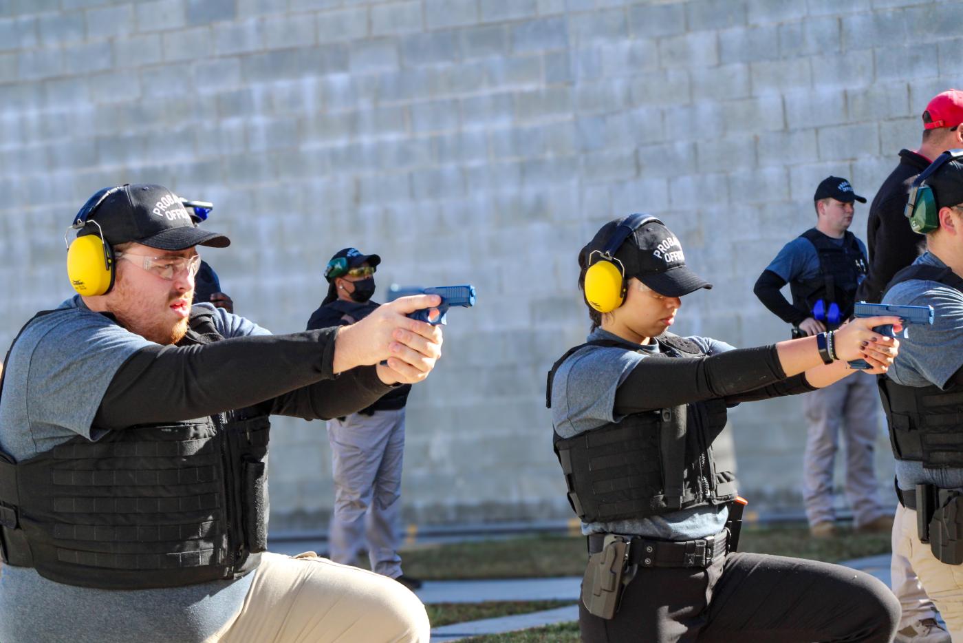 Multiple people stand in a line wearing yellow ear covers. Individuals are holding blue guns.