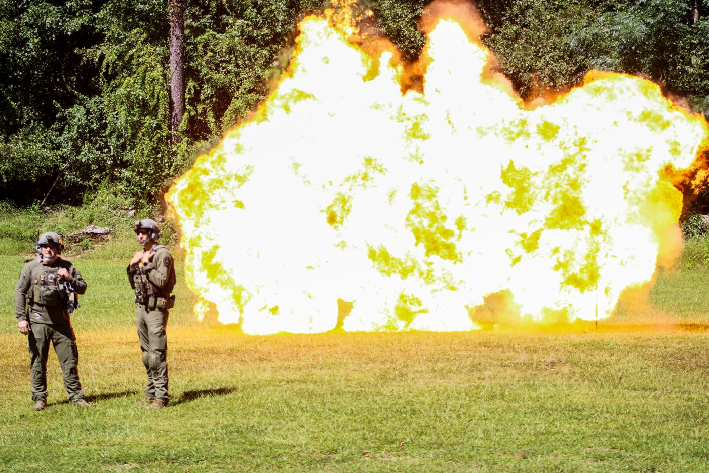 Two men in tactical gear stand in the foreground outdoors. In the background a large explosion erupts. 