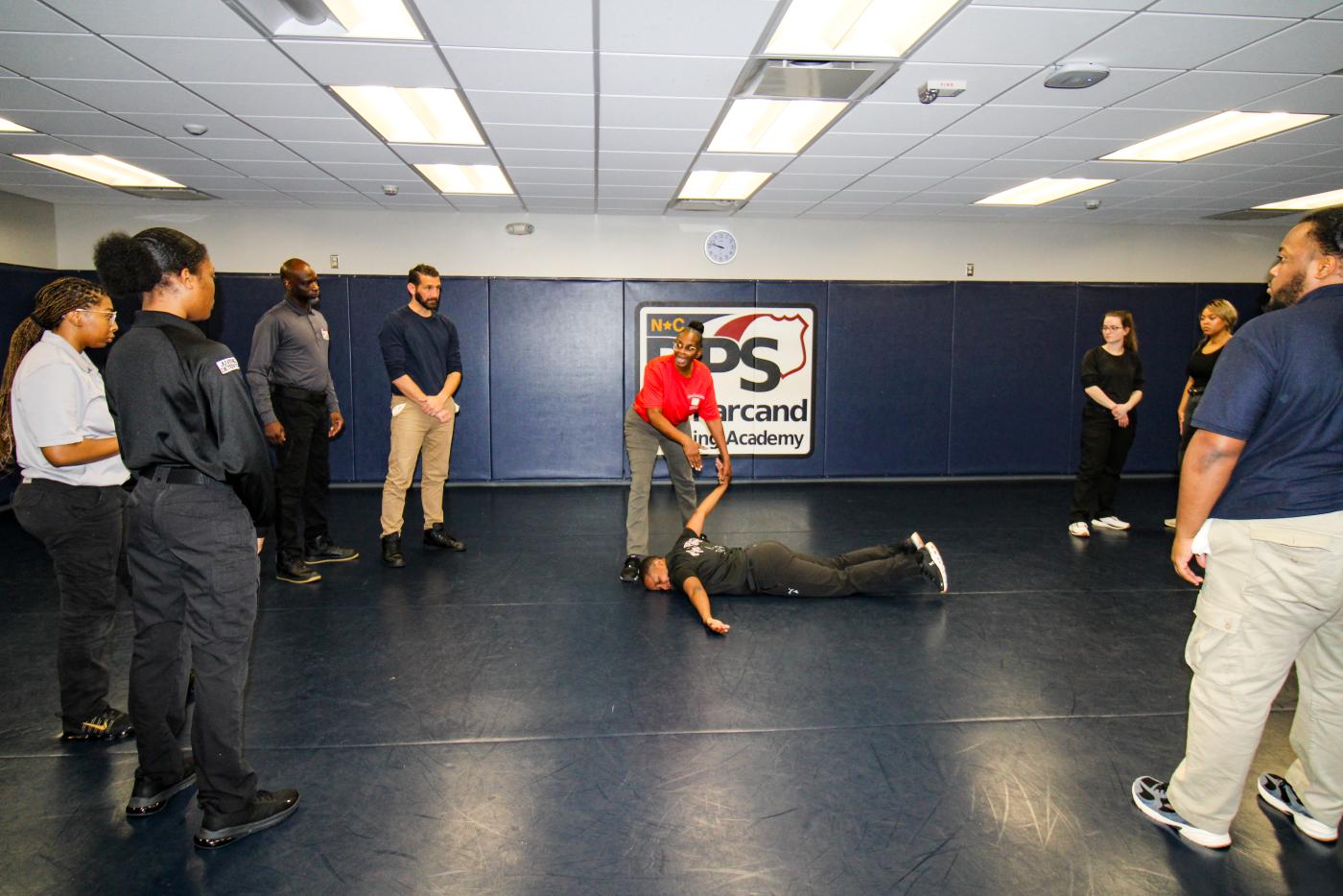 Group of people in a room with mats placed on floor and walls. A person lays on the floor in the middle with an instructor teaching a technique on them.