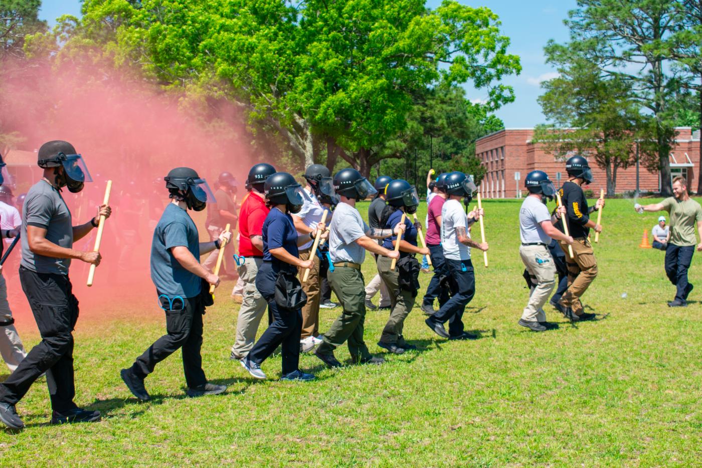 People walking in a line wearing helmets and masks and carrying batons. A red mist is in the background.