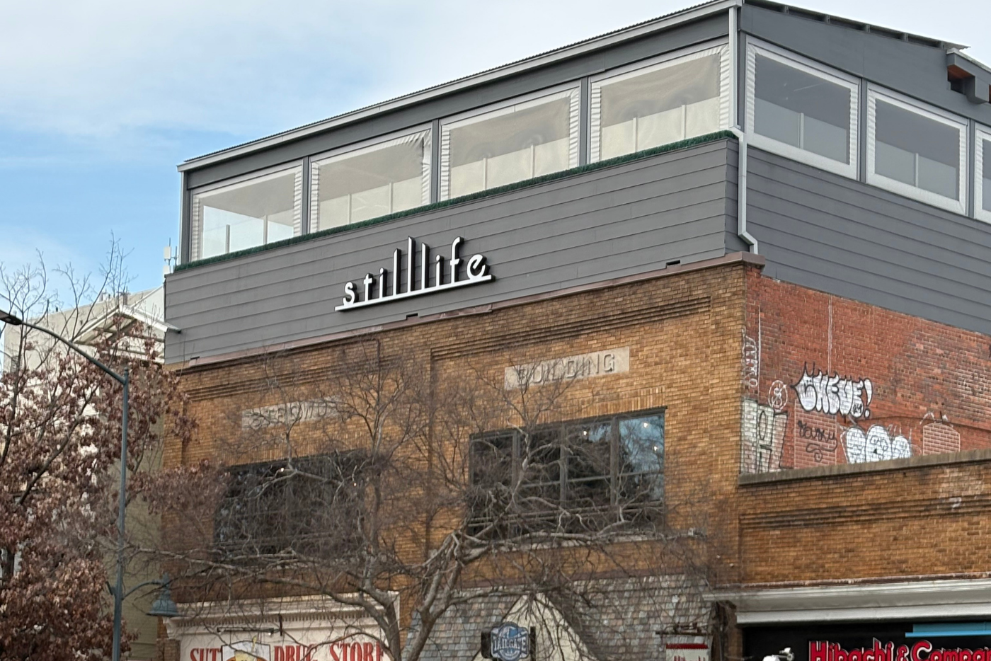Red brick building with grey siding with a business sign that displays the words still life on it