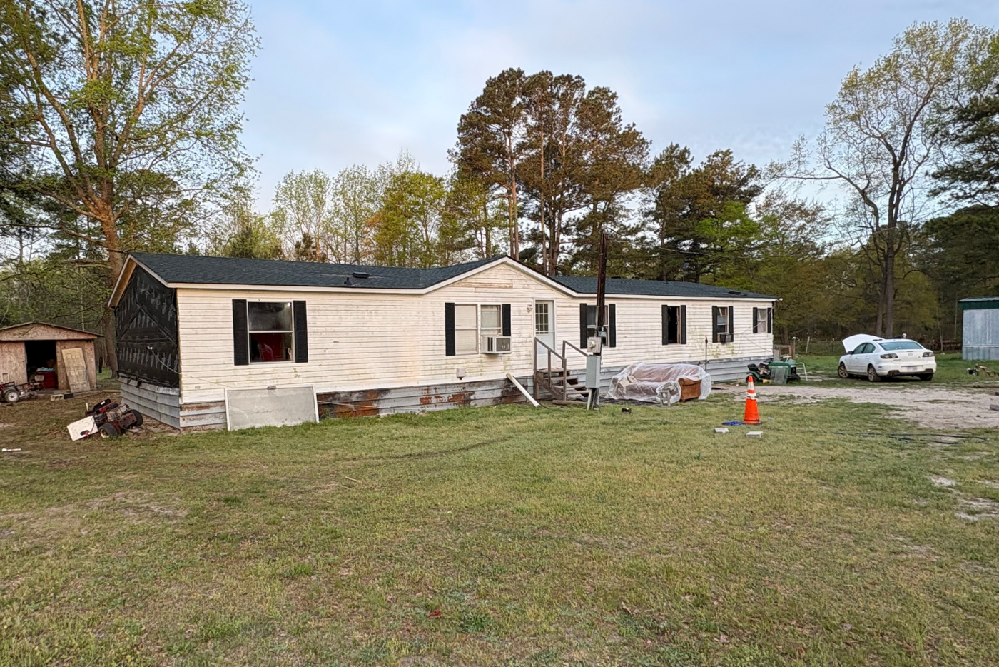 White mobile home with white mazda car in front with hood open. Brown couch sitting in front yard and a trash pile on the left side of the house where siding is missing 