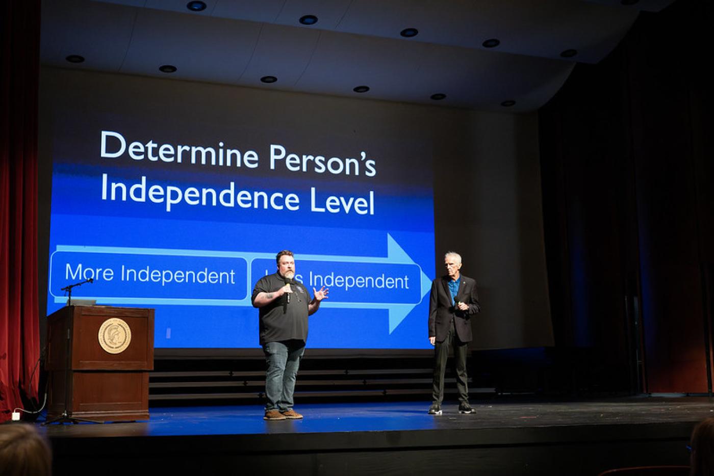 Two individuals speaking on a stage. A PowerPoint slide behind them is titled, "Determine Person's Independence Level."