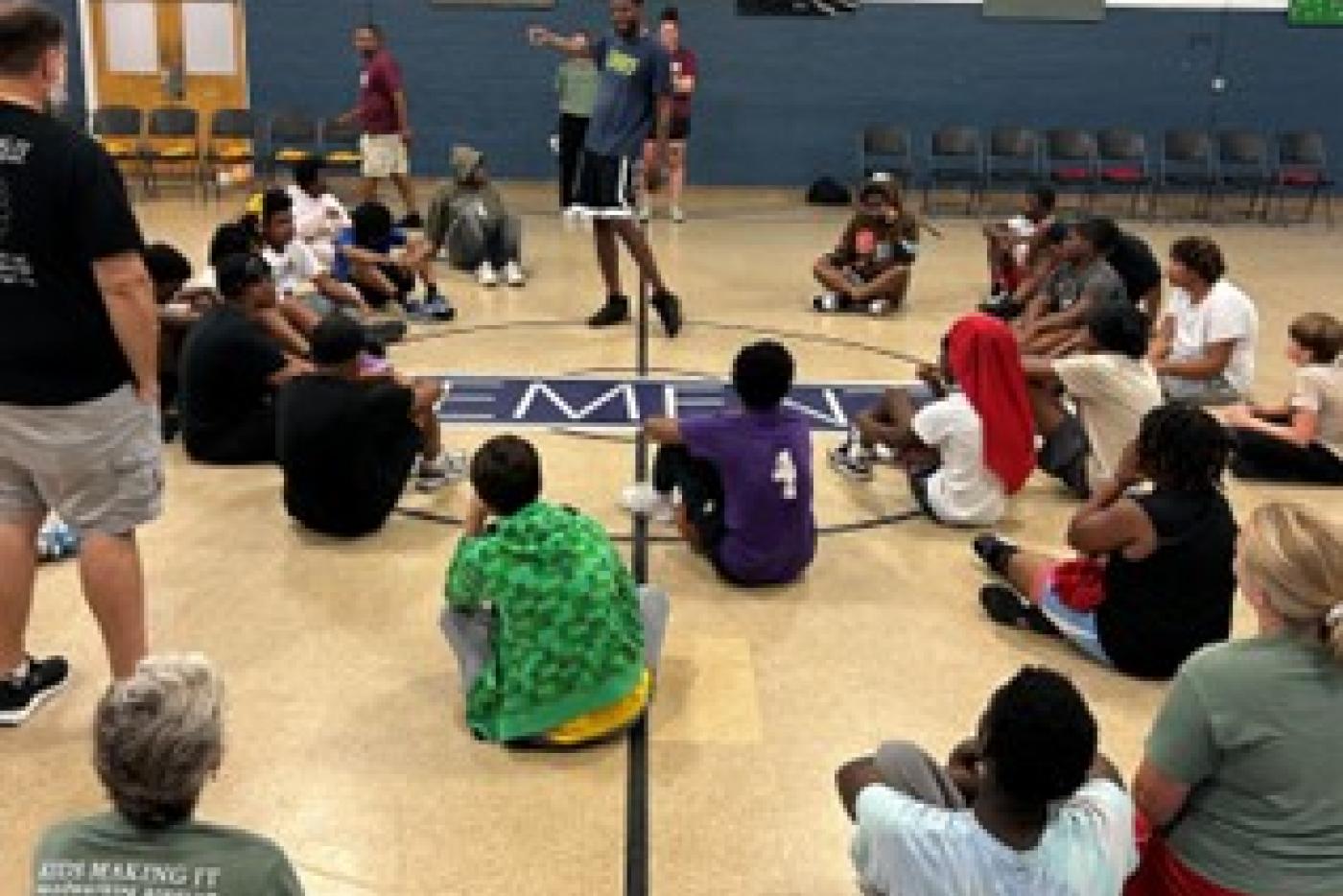 Group of kids sitting on floor of a gym listening to an adult speaker