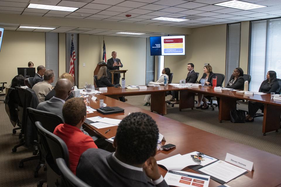 Multiple people sit at connected meeting tables. A male in a suit stands at the front of the room at a podium.