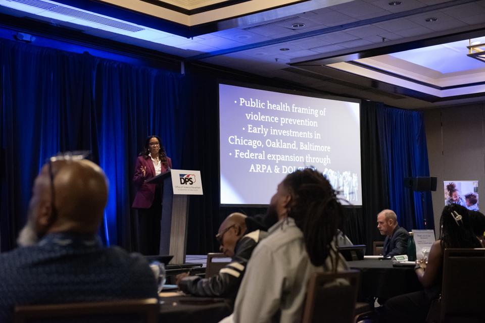 A speaker stands at a podium next to a large screen displaying a presentation slide.