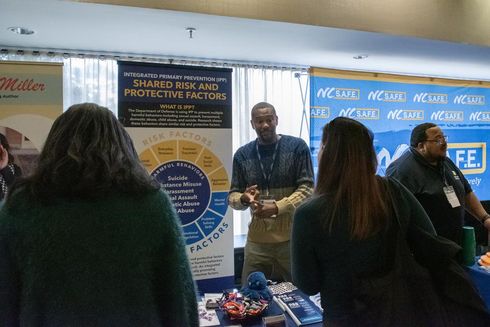 A group of people stands at an information booth with banners displaying "Integrated Primary Prevention" and "NC SAFE."