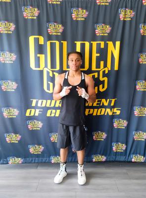 Young boxer stands in front of a “Golden Gloves Tournament of Champions” banner, wearing athletic gear with hands wrapped