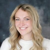 Woman with long blonde hair, wearing a white sweater, standing against a gray colored background