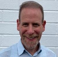Man with short hair and beard, wearing a blue collared button-down shirt with a white background