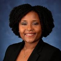 Woman with short black hair, wearing a black suit, sitting against a blue background