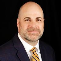 Man with gray beard wearing a dark suit, white shirt, and striped tie, sitting against a black background