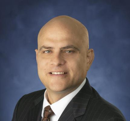 Matthew Rebuck in black suit in front of blue background.