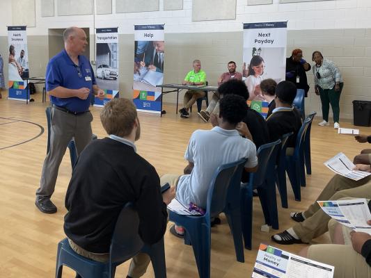 A person stands addressing a seated group youth in a gymnasium.